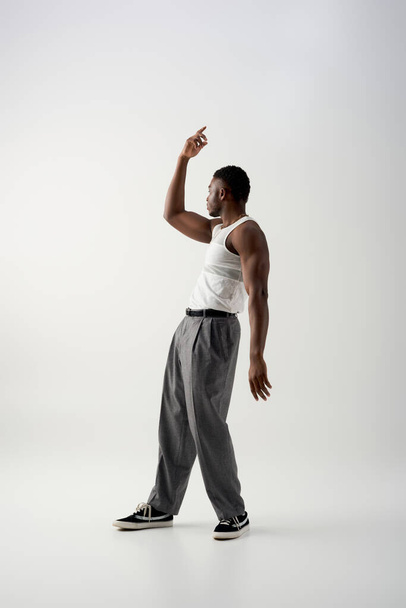 Widok z boku muskularny młody Afroamerykanin człowiek w koszulce bez rękawów i spodnie patrząc z boku na szarym tle, współczesna sesja featuring casual strój  - Zdjęcie, obraz