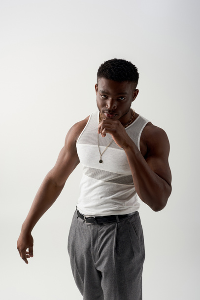 Confident young afroamerican man in tank top and pants looking at camera while standing isolated on grey, contemporary shoot featuring stylish attire, fashion design  - Фото, зображення