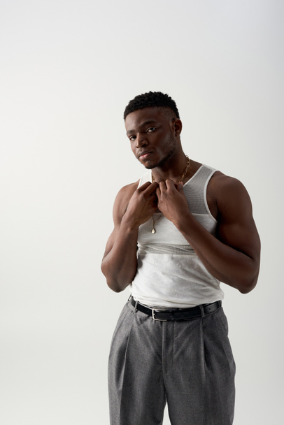 Muscular and young african american man in sleeveless t-shirt and pants touching necklace and looking at camera isolated on grey, contemporary shoot featuring casual attire - Foto, immagini