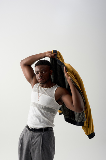 Homem afroamericano jovem confiante no topo do tanque usando jaqueta de bombardeiro e de pé isolado em cinza, tiro contemporâneo com traje elegante, muscular, fashion-forward  - Foto, Imagem