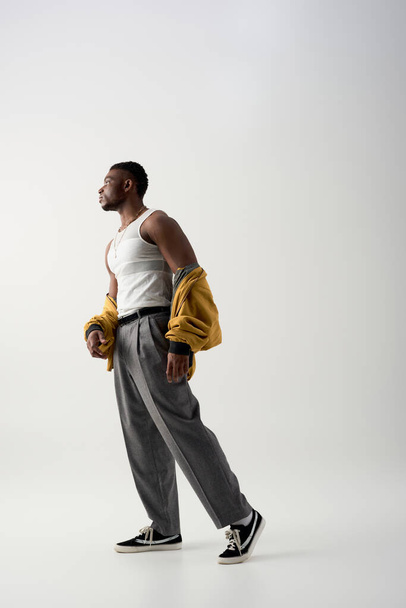 Oldalnézet izmos afro-amerikai férfi bomber kabát és ujjatlan póló álló szürke háttér, kortárs fotózás jellemző elegáns öltözék, divat nyilatkozat  - Fotó, kép