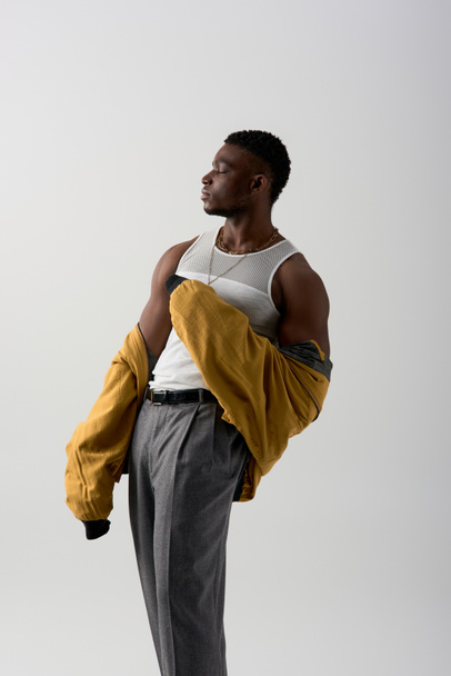 Side view of good looking afroamerican model in bomber jacket and sleeveless t-shirt posing isolated on grey, contemporary shoot featuring stylish attire, muscular - Photo, Image