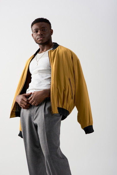 Magabiztos fiatal afro-amerikai férfi bombázó kabát és nadrág pózol és néz kamera elszigetelt szürke, kortárs fotózás jellemző stílusos öltözék, divat nyilatkozat  - Fotó, kép