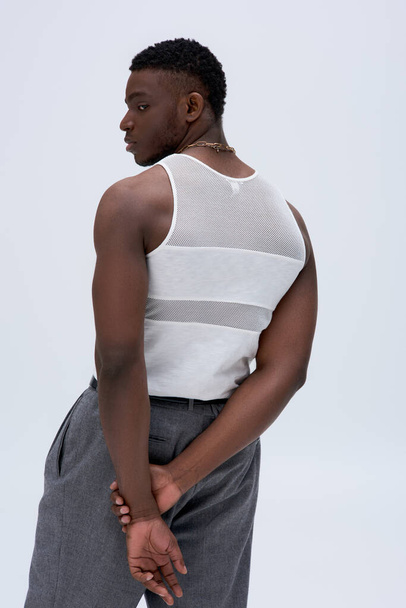 Oldalnézet a sportos fiatal afro-amerikai férfi a tartály felső és nadrág néz félre, és pózol magabiztosan elegáns és divatos ruhát elszigetelt szürke, jóképű    - Fotó, kép