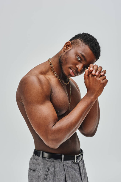 Muscular and good looking afroamerican man in golden necklaces and pants looking at camera while standing isolated on grey, confident and modern pose, fashion shoot - Foto, afbeelding
