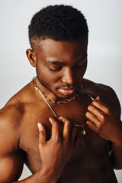 Young and shirtless african american male model touching golden necklaces and standing isolated on grey, confident and modern pose, fashion shoot - Фото, зображення