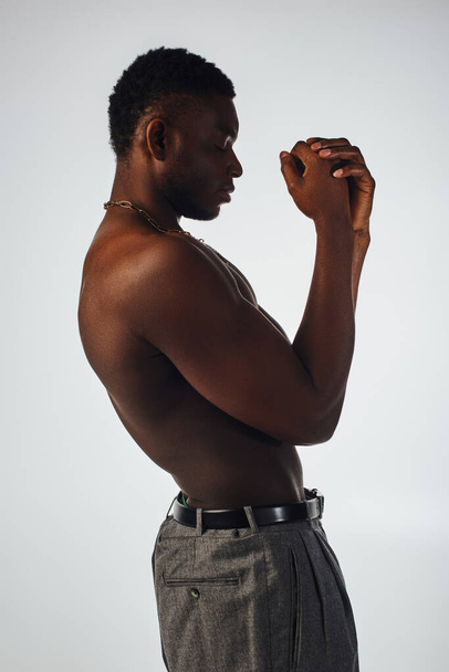 Zijaanzicht van ontspannen en shirtloze Afro-Amerikaanse man in kettingen en broeken geïsoleerd op grijze, zelfverzekerde en moderne pose, fashion shoot, gespierd model  - Foto, afbeelding