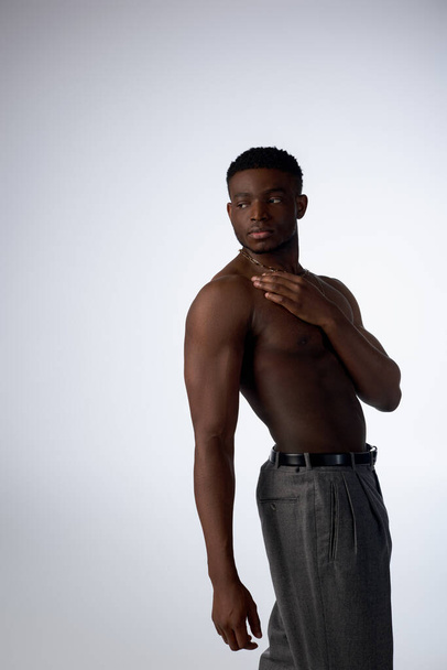 Shirtless and young afroamerican man in golden necklaces and pants standing in shadow isolated on grey, confident and modern pose, fashion shoot, muscular model  - Foto, afbeelding