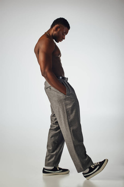 Vista lateral do homem afroamericano muscular em colares dourados e calças de mão no bolso em fundo cinza, pose confiante e moderna, tiro de moda, modelo sem camisa  - Foto, Imagem