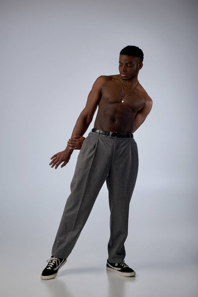 Plná délka svalnatého afroamerického modelu ve zlatých náhrdelnících, kalhotách a teniskách stojící na šedém pozadí, sebevědomá a moderní póza, módní natáčení, muž bez trička - Fotografie, Obrázek