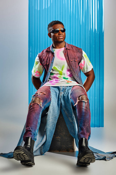 Full length of stylish african american model in sunglasses, denim vest and ripped jeans sitting on stone on grey with blue polycarbonate sheet at background, fashion shoot, sustainable fashion  - Photo, Image