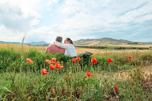 Пара средних лет, сидящая на поляне из маков и поле из золотой пшеницы в жарком летнем солнце и голубом небе с белыми облаками на фоне горных холмов. - Фото, изображение