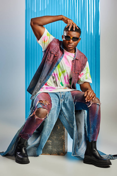 Stylish afroamerican man in sunglasses, denim vest and colorful t-shirt touching head while sitting on stone on grey with blue polycarbonate sheet at background, fashion shoot, sustainable fashion  - 写真・画像