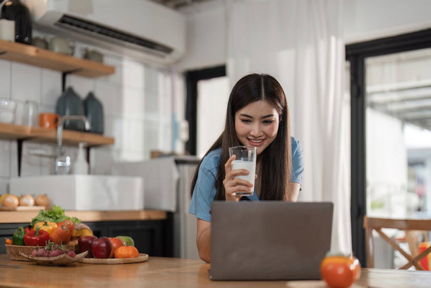 Belle jeune femme avec ordinateur portable boire du lait dans la cuisine
. - Photo, image