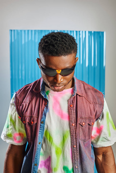 Portrait of young and stylish afroamerican man with trendy hairstyle wearing sunglasses, denim vest and colorful t-shirt on grey with blue polycarbonate sheet at background, sustainable fashion - Photo, Image