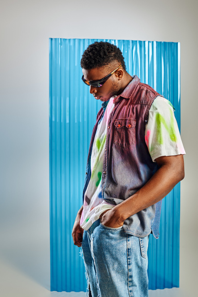 Trendy young afroamerican man in sunglasses, colorful t-shirt and denim vest holding hand in pocket of ripped jeans on grey with blue polycarbonate sheet at background, DIY clothing - Photo, Image