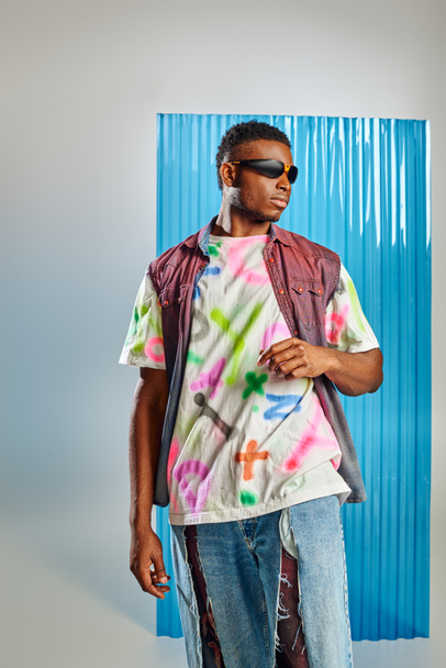Módní mladý afroamerický model ve slunečních brýlích, barevné džínové vestě a roztržených džínách stojící na šedé s modrým polykarbonátovým prostěradlem na pozadí, udržitelná móda, domácí oblečení - Fotografie, Obrázek