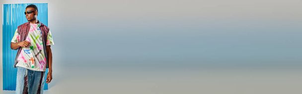 Jeune modèle afroaméricain en gilet en denim coloré, lunettes de soleil et élégant jean déchiré debout sur gris avec feuille de polycarbonate bleu à l'arrière-plan, tournage de mode, bannière, vêtements DIY - Photo, image