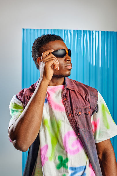 Portrait of trendy afroamerican man touching sunglasses while wearing denim vest and colorful t-shirt on grey with blue polycarbonate sheet at background, sustainable fashion, DIY clothing - Photo, Image