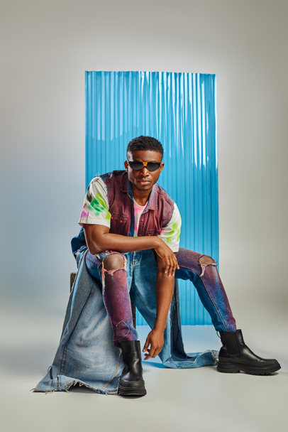 afroamerykański model w okularach przeciwsłonecznych, kolorowa kamizelka jeansowa i współczesne podarte dżinsy siedzące na kamieniu na szarości z niebieskim prześcieradłem poliwęglanowym na tle, zrównoważona moda, odzież DIY - Zdjęcie, obraz