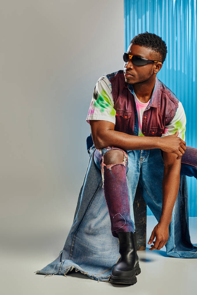 Confident afroamerican man in sunglasses, denim vest and trendy ripped jeans sitting on stone on grey with blue polycarbonate sheet at background, sustainable fashion, DIY clothing - Photo, Image