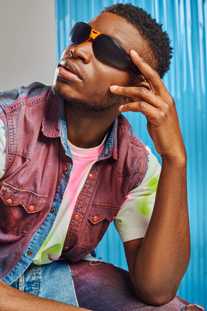 Πορτρέτο του κομψού afroamerican μοντέλο σε γυαλιά ηλίου πολύχρωμο γιλέκο denim και t-shirt ποζάρουν σε γκρι με μπλε πολυανθρακικό φύλλο στο παρασκήνιο, φωτογράφιση μόδας, DIY ρούχα, βιώσιμο τρόπο ζωής  - Φωτογραφία, εικόνα