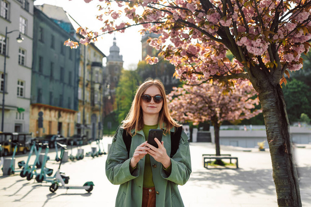 Ropa elegante chica rubia turista viaja en busca de direcciones en el teléfono inteligente mientras viaja a través del casco antiguo de Cracovia, cerca del Castillo Real de Wawel en primavera, Polonia. Primavera Flores de cerezo - Foto, Imagen