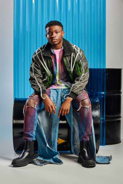 Trendikäs nuori afroamerikkalainen mies päällystakissa led-raidat ja revitty farkut istuu polttoaineen tynnyri ja katsot kameran lähellä sininen polykarbonaatti arkki harmaalla pohjalla, DIY vaatteet  - Valokuva, kuva
