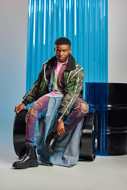 Modelo afroamericano joven confiado con peinado de moda que usa chaqueta de traje con rayas led y jeans rasgados mientras está sentado en el barril de combustible cerca de la hoja de policarbonato azul en gris, ropa de bricolaje  - Foto, Imagen