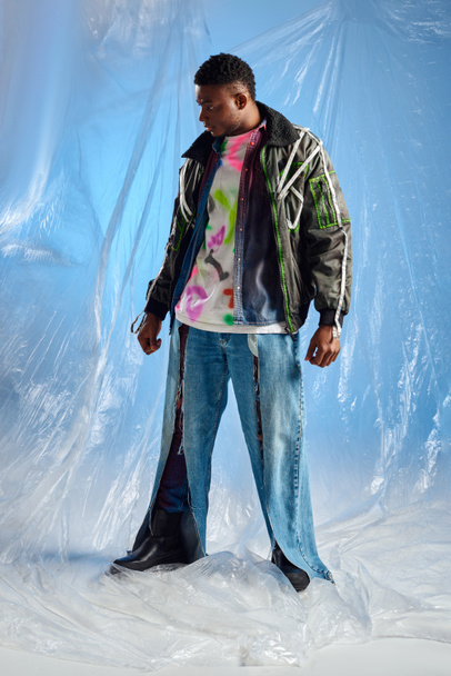 Full length of stylish afroamerican male model in outwear jacket with led stripes and ripped jeans looking away while standing on glossy cellophane on blue background, urban outfit, DIY clothing  - Фото, зображення