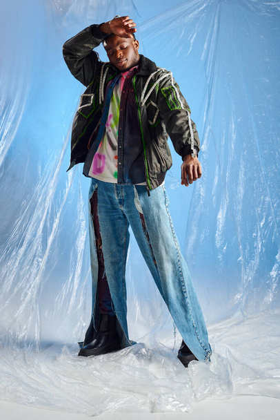 Boa aparência jovem modelo afroamericano em jaqueta de roupas com listras led e jeans rasgados tocando a testa e em pé sobre celofane brilhante no fundo azul, roupa urbana e roupas DIY  - Foto, Imagem