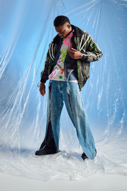 Hombre afroamericano joven guapo en chaqueta de outwear con rayas led mirando vaqueros rasgados de moda de pie en celofán brillante sobre fondo azul, traje urbano, ropa de bricolaje, estilo de vida sostenible  - Foto, Imagen