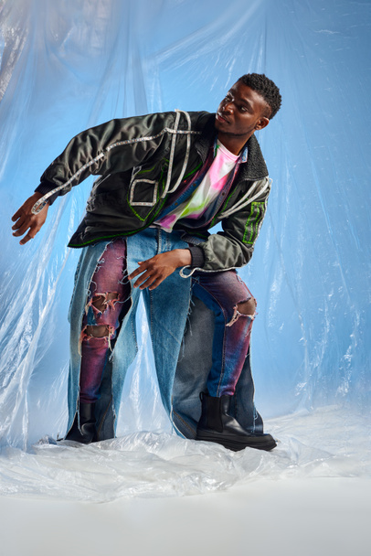 Elegante modelo afroamericano joven en chaqueta de outwear con rayas led y jeans rasgados mirando hacia otro lado y posando sobre celofán brillante sobre fondo azul, atuendo urbano y estilo de vida sostenible  - Foto, Imagen