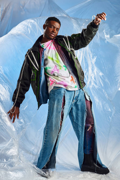 In voller Länge selbstbewusster afroamerikanischer Mann in Outwear-Jacke mit Bleistreifen und zerrissenen Jeans, der in die Kamera schaut, in der Nähe von glänzendem Zellophan auf blauem Hintergrund, moderne Pose, nachhaltiger Lebensstil  - Foto, Bild
