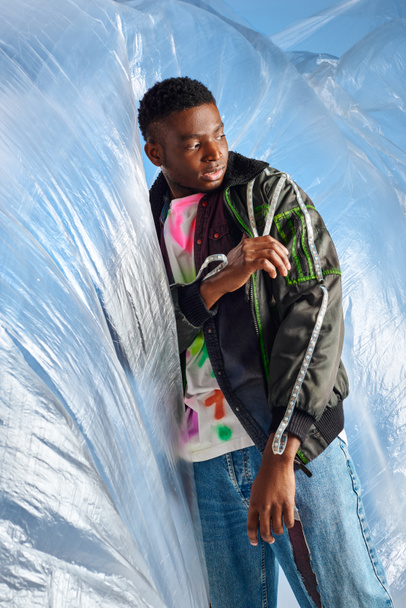 Modelo masculino afroamericano joven de moda en jeans rasgados tocando chaqueta de outwear con rayas led cerca de celofán brillante sobre fondo azul, traje urbano, ropa de bricolaje, estilo de vida sostenible  - Foto, imagen