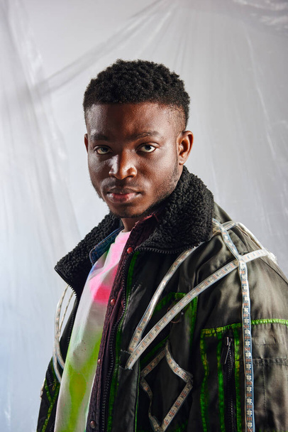 Portrait of young afroamerican model in outwear jacket with led stripes looking at camera near glossy cellophane on grey background, urban outfit and modern pose, creative expression, DIY clothing  - 写真・画像
