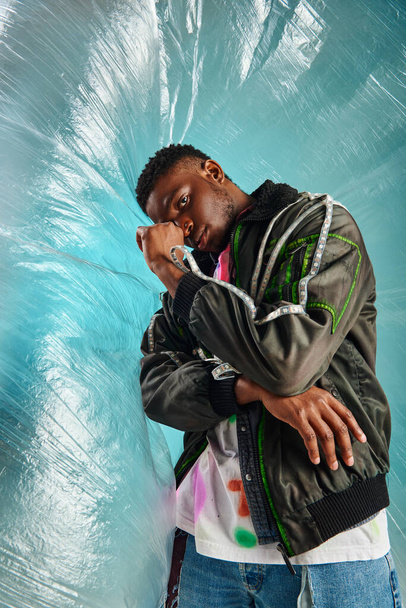 Confident young african american model in outwear jacket with led stripes and shriffed jeans posing near glossy cellophane on turquoise background, urban outfit, δημιουργική έκφραση, DIY ρούχα  - Φωτογραφία, εικόνα