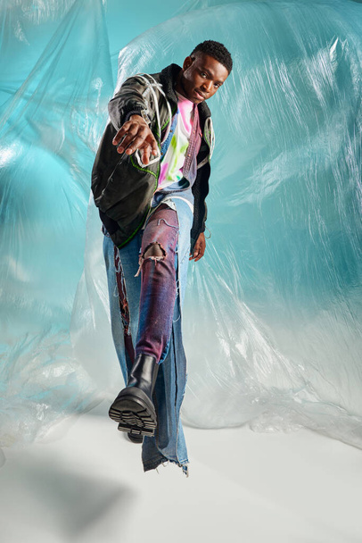 Comprimento total do jovem modelo afroamericano na moda em jaqueta de roupas e jeans rasgados olhando para a câmera e andando perto de celofane brilhante em fundo turquesa, expressão criativa, roupas DIY  - Foto, Imagem