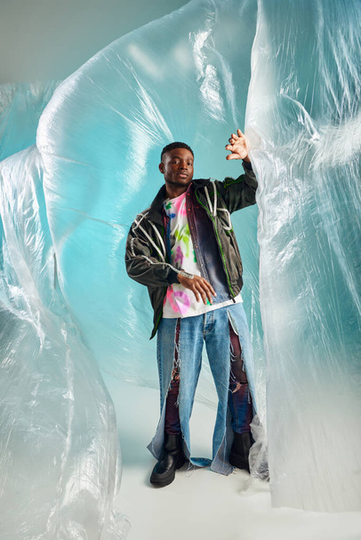 Full length of trendy young african american man in ripped jeans and outwear jacket looking away near glossy cellophane on turquoise background, urban outfit, creative expression, DIY clothing  - 写真・画像