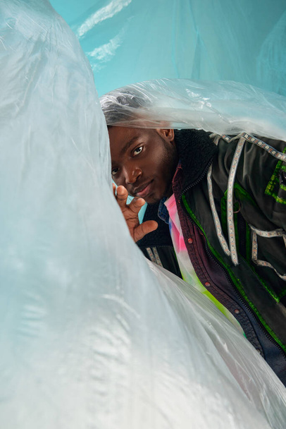 Fashionable young african american man in outwear jacket with led stripes standing near glossy cellophane on turquoise background, urban outfit and modern pose, creative expression, DIY clothing  - 写真・画像