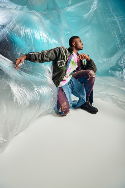 Full length of young afroamerican model in ripped jeans and outwear jacket with led stripes looking away near cellophane on turquoise background, modern pose, creative expression, DIY clothing  - Fotó, kép
