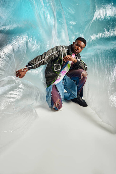 Fashionable and confident young african american model in outwear jacket and ripped jeans looking at camera near cellophane on turquoise background, creative expression, DIY clothing  - Фото, зображення