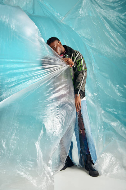 african american man in ripped jeans and outwear jacket with led stripes covering face with cellophane on turquoise background, urban outfit and modern pose, creative expression, DIY clothing  - Photo, Image