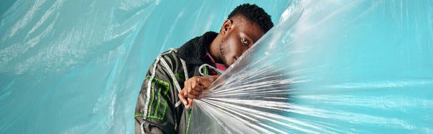 Young and trendy afroamerican model in outwear jacket and led stripes covering face with cellophane on turquoise background, urban outfit and modern pose, banner, creative expression, DIY clothing  - Photo, Image