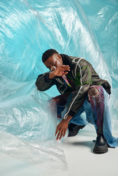 creative expression, DIY clothing, young african american model in outwear jacket with led stripes and trendy jeans posing and covering face near cellophane on turquoise background  - Foto, imagen