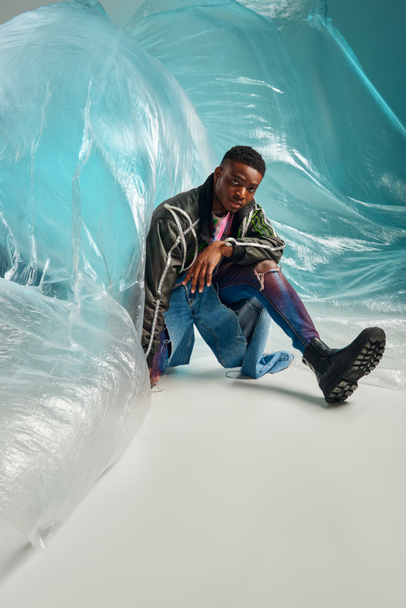 Young afroamerican model in outwear jacket with led stripes and ripped jeans sitting near glossy cellophane on turquoise background, urban outfit and modern pose, creative expression, DIY clothing  - Foto, Imagen