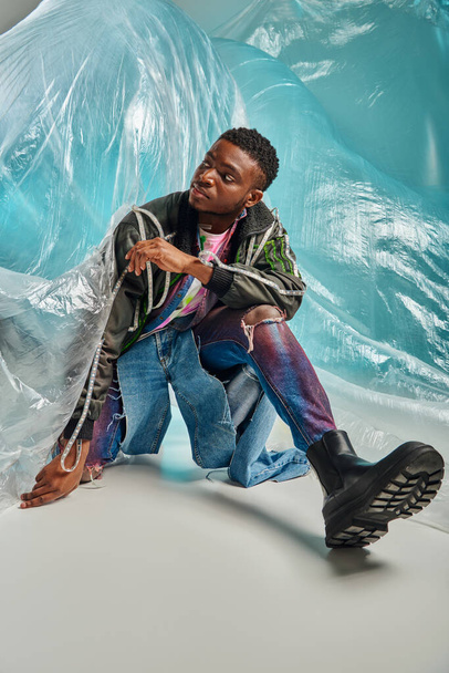 Modelo masculino afroamericano de moda en jeans rasgados y chaqueta de outwear con rayas led mirando hacia otro lado y posando cerca de celofán sobre fondo turquesa, expresión creativa, ropa de bricolaje  - Foto, imagen