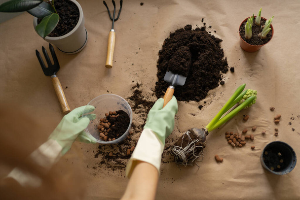 A kesztyűs női kezek talajt öntenek, és vízelvezetést tesznek a spatulával rendelkező növények átültetésére szolgáló edénybe. Tavaszi szobanövények gondozása és újratelepítése - Fotó, kép