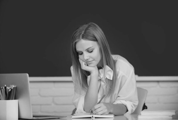 Portrait of young female college student studying in classroom on class with blackboard background. Student education concept. Female student thinking about coursework in university - Photo, image