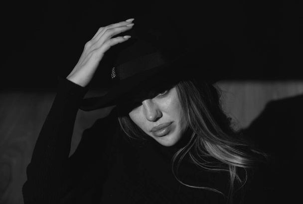 Girl in fashion hat. Young woman posing at studio over black background. Light and shadow. Fashion portrait of beautiful woman with dark light on face. Hidden eyes with wide broad brim hat - Fotó, kép